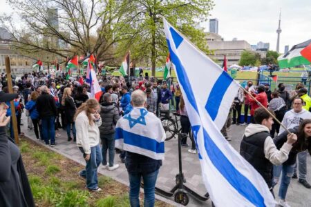 Pro-Israel supporters holding Israeli flags walks through crowd as pro-Palestinian and pro-Israeli protesters passionately express their views, with intense exchanges outside the student encampment occupying King's College Circle at the University of Toronto.
